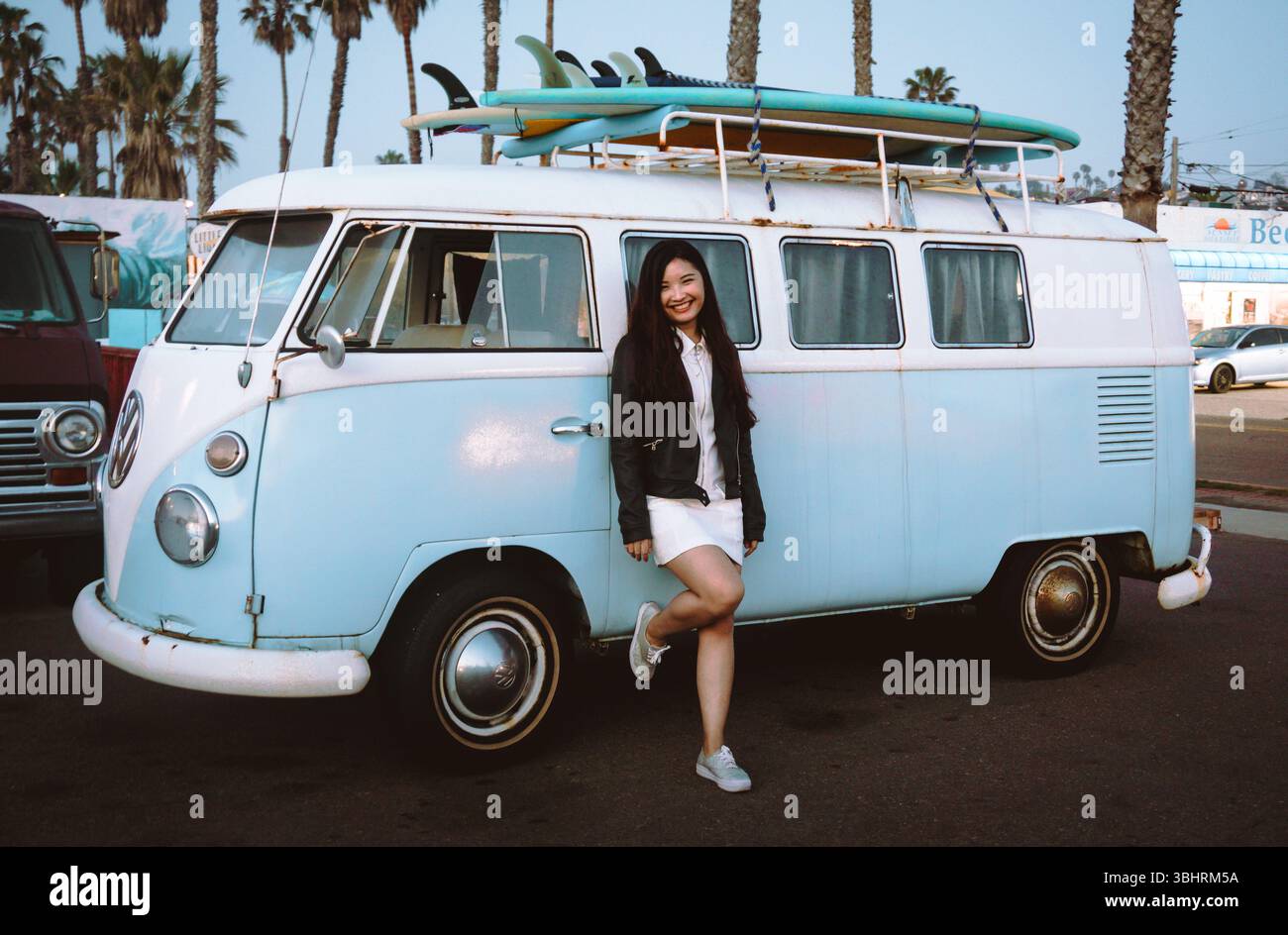 Un pulmino Volkswagen d'epoca con una tavola da surf in cima si trova sotto le palme, catturando l'avventura costiera e le vibrazioni del viaggio retrò. Foto Stock