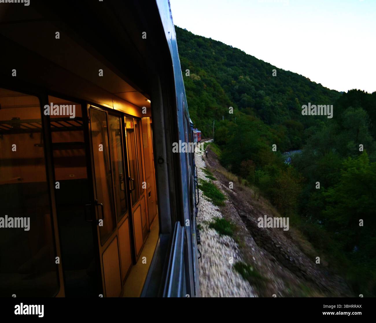 Treno bulgaro sulla strada da Bucarest a Sofia passando attraverso la foresta panoramica e le colline durante il crepuscolo Foto Stock