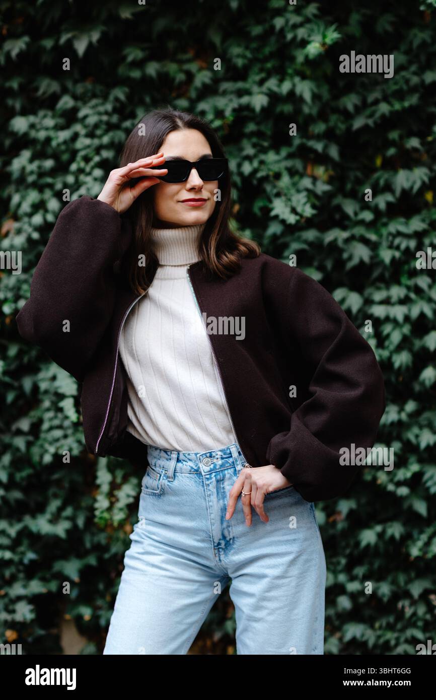 Giovane donna elegante che indossa occhiali da sole, bomber, maglione dolcevita e jeans in posa davanti a una parete ricoperta di edera Foto Stock