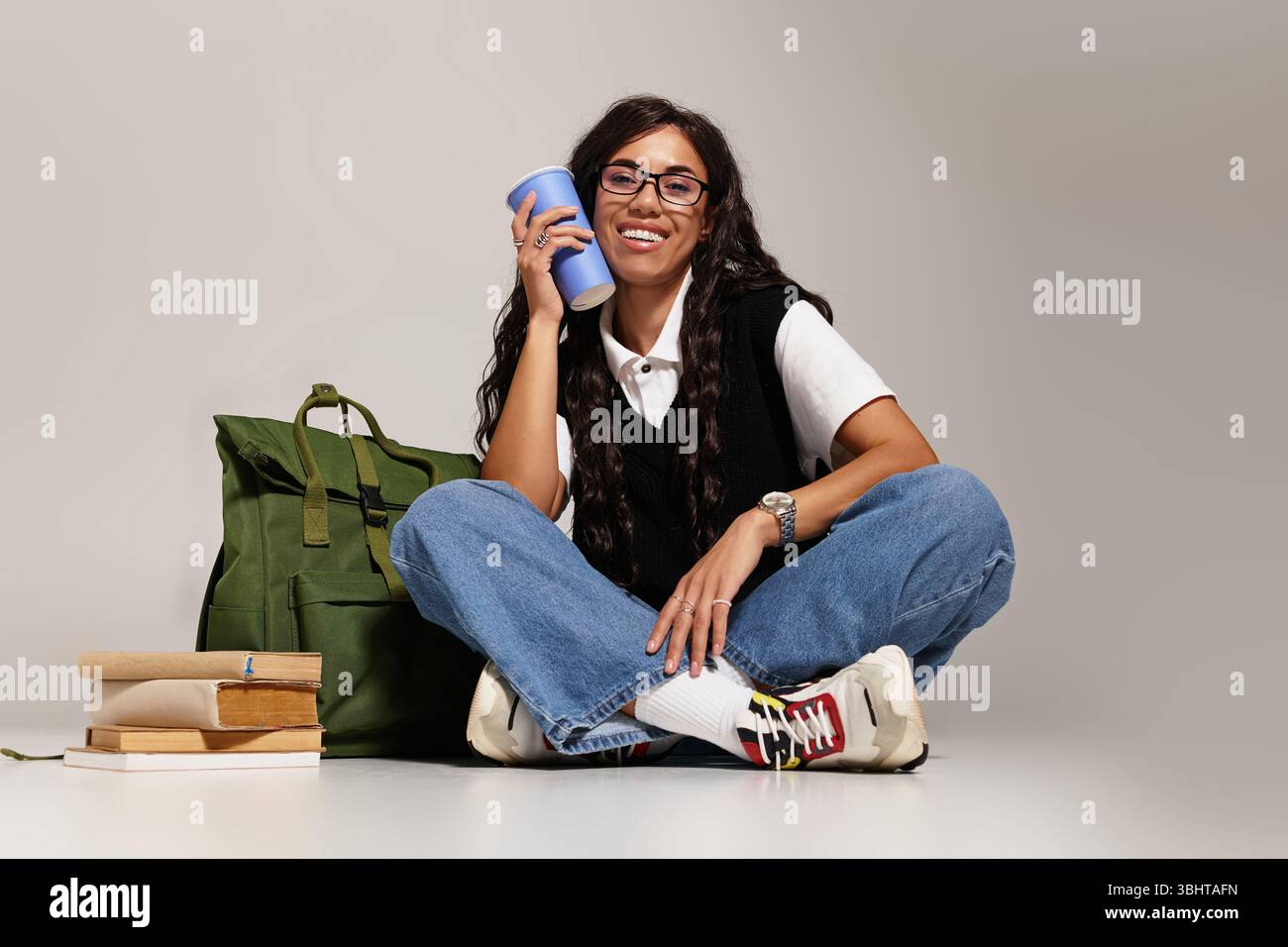 Uno studente allegro gode di un momento di relax con libri, uno zaino e un drink in mano. Foto Stock