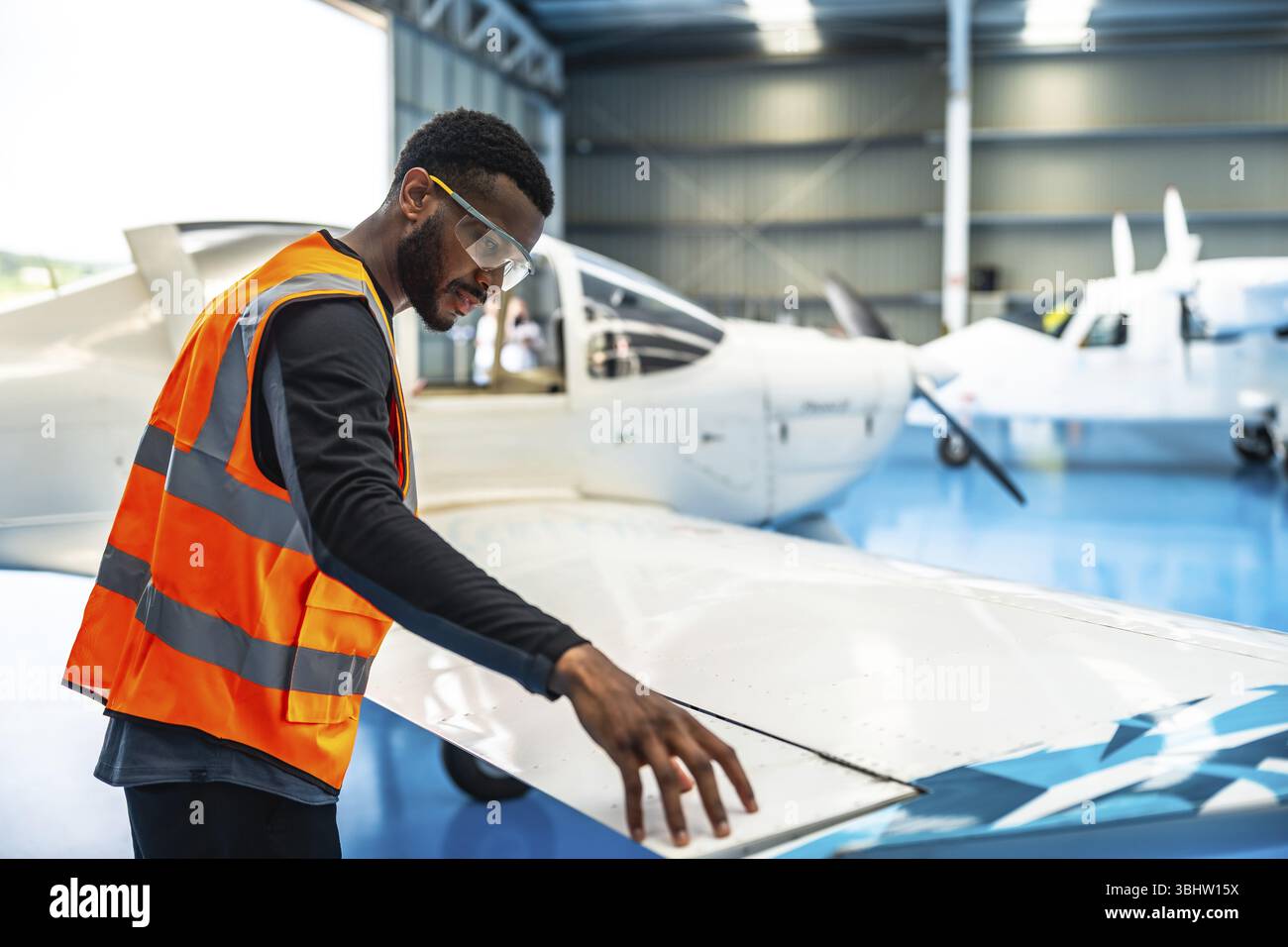 Un tecnico addetto alla manutenzione degli aeromobili che indossa occhiali di sicurezza e un giubbotto ad alta visibilità controlla l'ala di un piccolo aereo passeggeri all'interno di un hangar Foto Stock