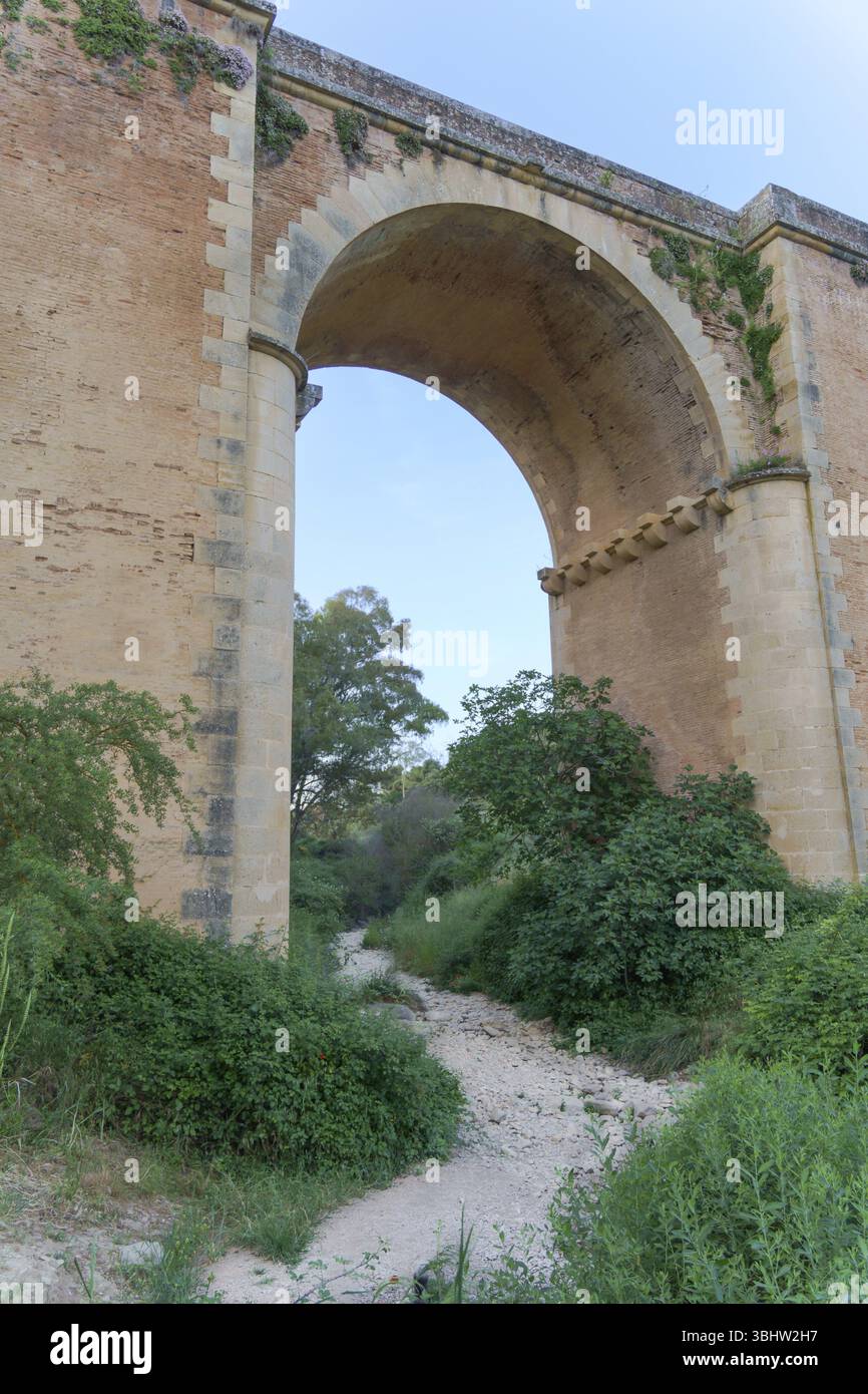 Ronda, malaga, spagna letto di fiume secco con rocce e vegetazione che passa sotto un vecchio ponte di mattoni con un grande arco Foto Stock