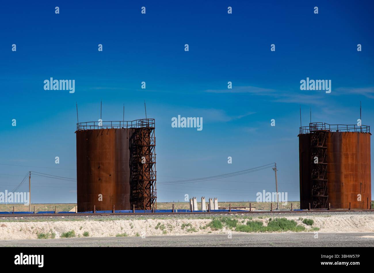 La foto mostra due grandi serbatoi di stoccaggio arrugginiti in un'area aperta sotto un cielo azzurro. Tra di loro ci sono poli di potenza ed elementi industriali Foto Stock