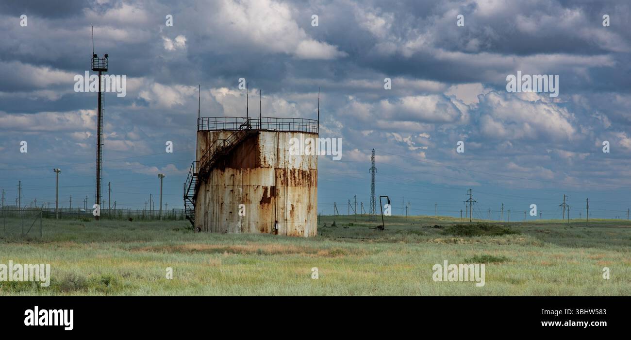 La foto mostra un serbatoio di stoccaggio dell'olio cilindrico arrugginito in un campo aperto. Attorno ad esso si trovano steppa, linee elettriche e una torre di guardia contro un dra Foto Stock