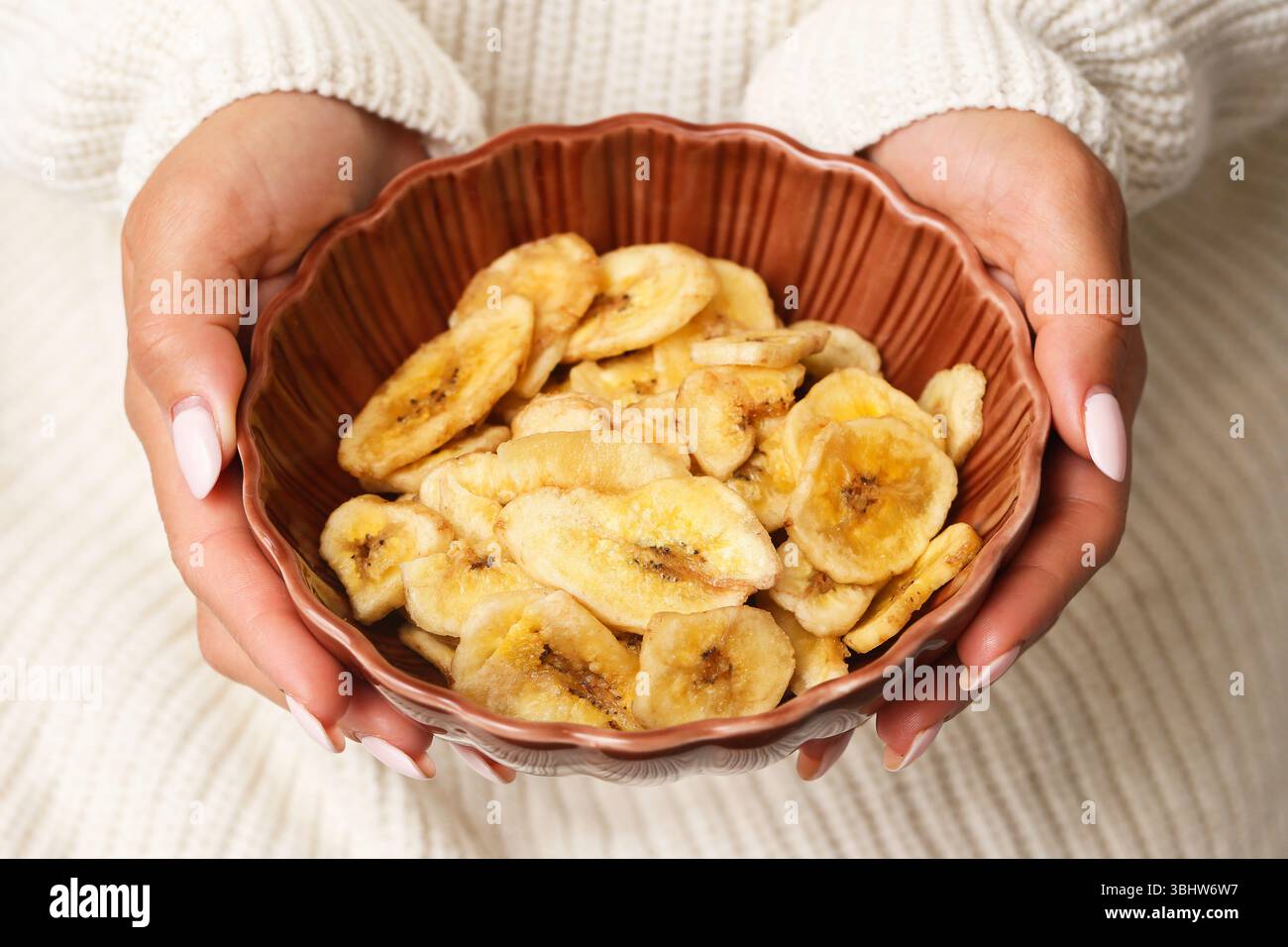 Gocce di banana essiccate in un recipiente di ceramica. Mani che tengono uno spuntino dolce. Frutta tropicale croccante. Sana alternativa alle caramelle. Consistenza naturale degli alimenti secchi. Foto Stock