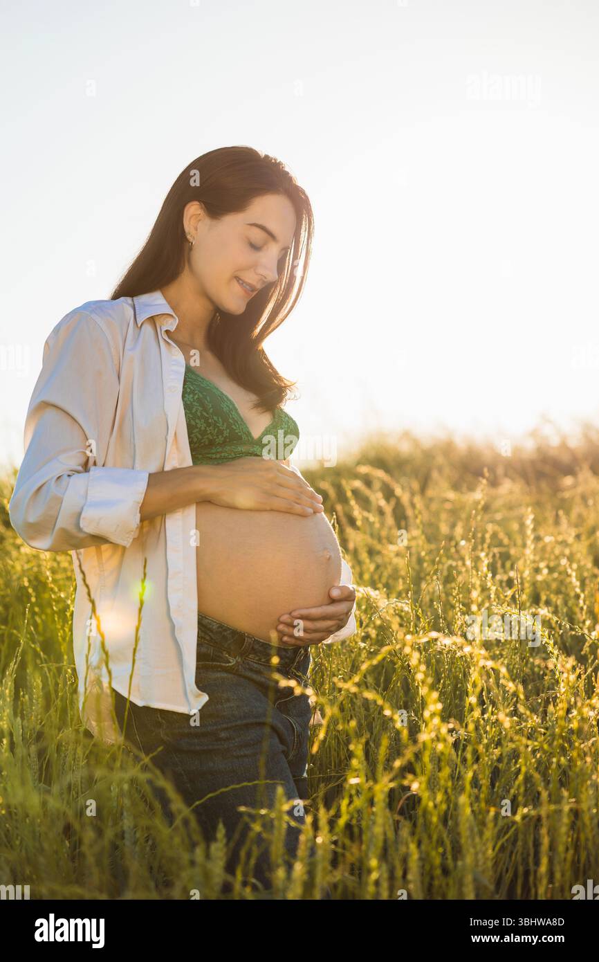 La mamma incinta si trova in un campo tranquillo al tramonto, indossando un reggiseno in pizzo verde e una camicia aperta, toccando delicatamente la pancia e godendosi il calore di Foto Stock