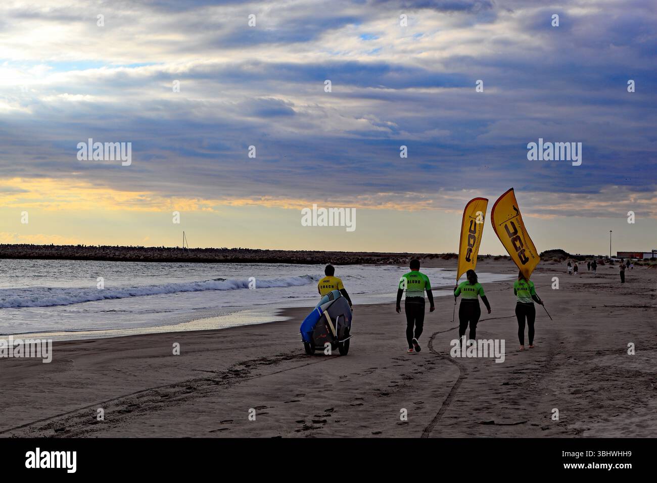Viana do Castelo, Portogallo - 2 marzo 2025: Un gruppo di surfisti che camminano lungo la spiaggia di Praia do Cabedelo al tramonto con tavole da surf e bandiere Foto Stock