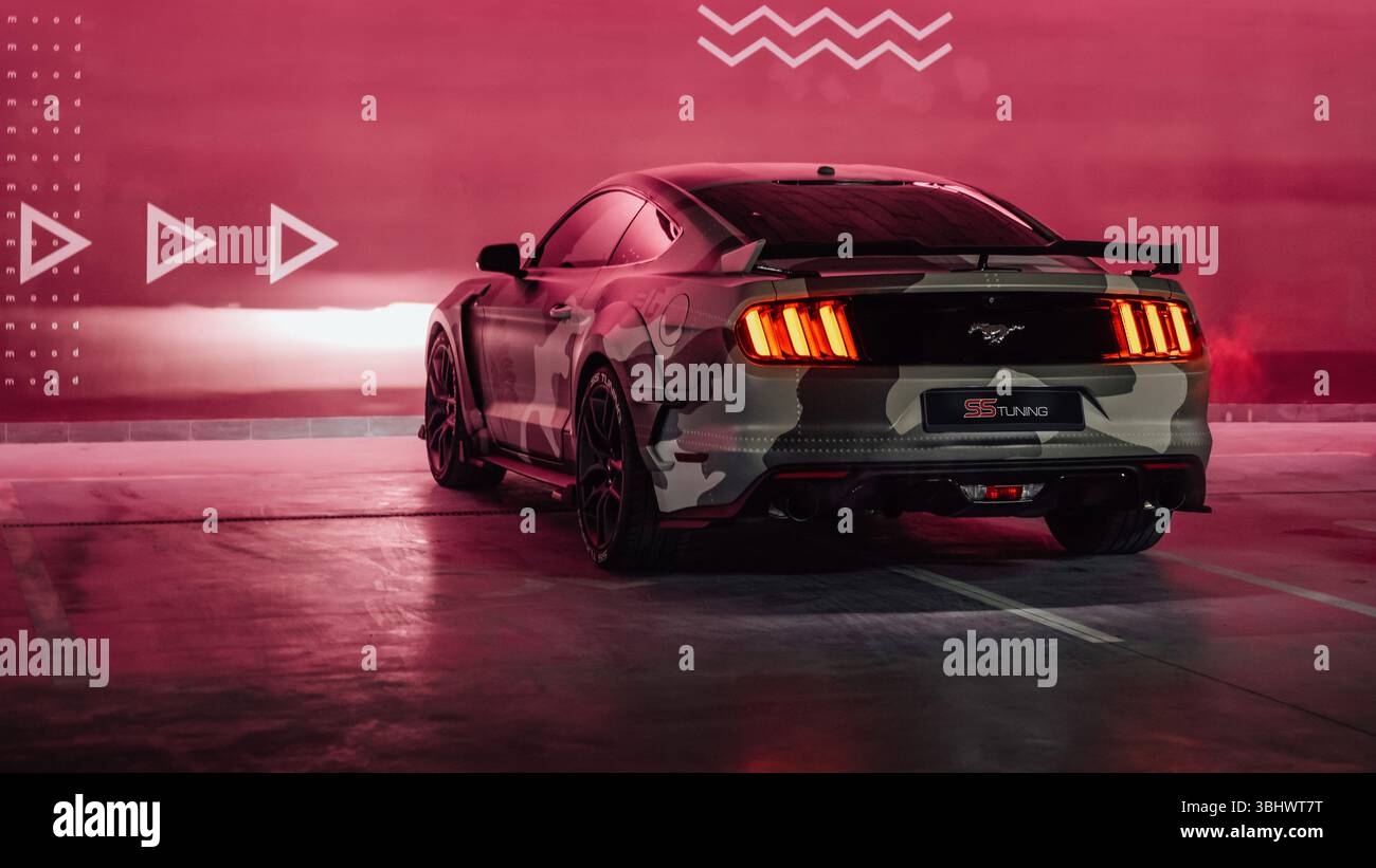 Ford Mustang beige camo nel garage. Vista posteriore di tre quarti del pony americano parcheggiato vicino alla parete marrone illuminata. Foto Stock