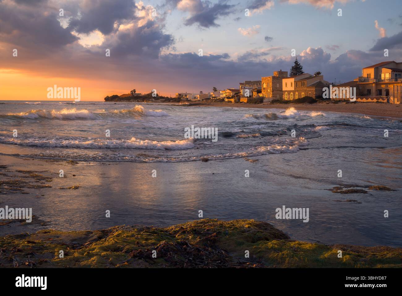 Le onde si infrangono dolcemente sulla spiaggia sabbiosa mentre il sole tramonta dietro le affascinanti case di Punta secca, offrendo un momento di tranquillità, la Sicilia, l'Italia Foto Stock