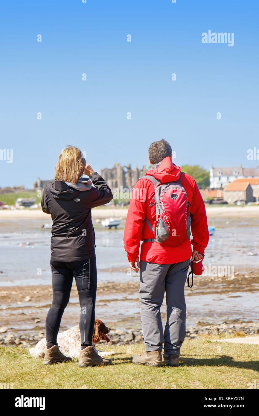 Due escursionisti con un cane in piedi su una costa erbosa del northumberland, che si affaccia su un villaggio di lindisfarne sull'isola Santa e sulla costa rocciosa sotto un cielo azzurro Foto Stock