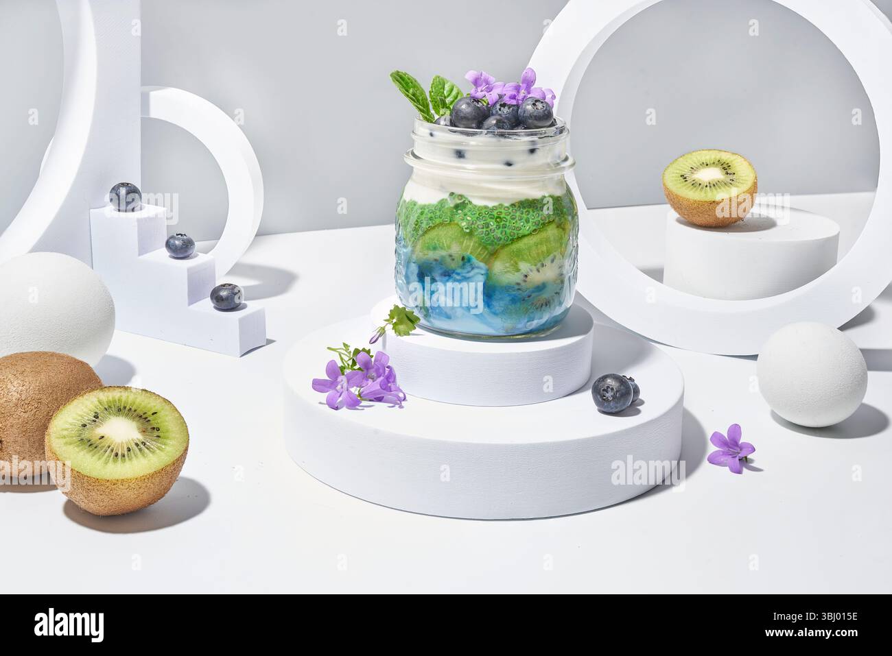 Un vivace parfait di yogurt in un barattolo con fette di kiwi fresche, mirtilli e artistiche tonalità verde e viola, presentato su uno sfondo bianco minimalista con wi-fi Foto Stock