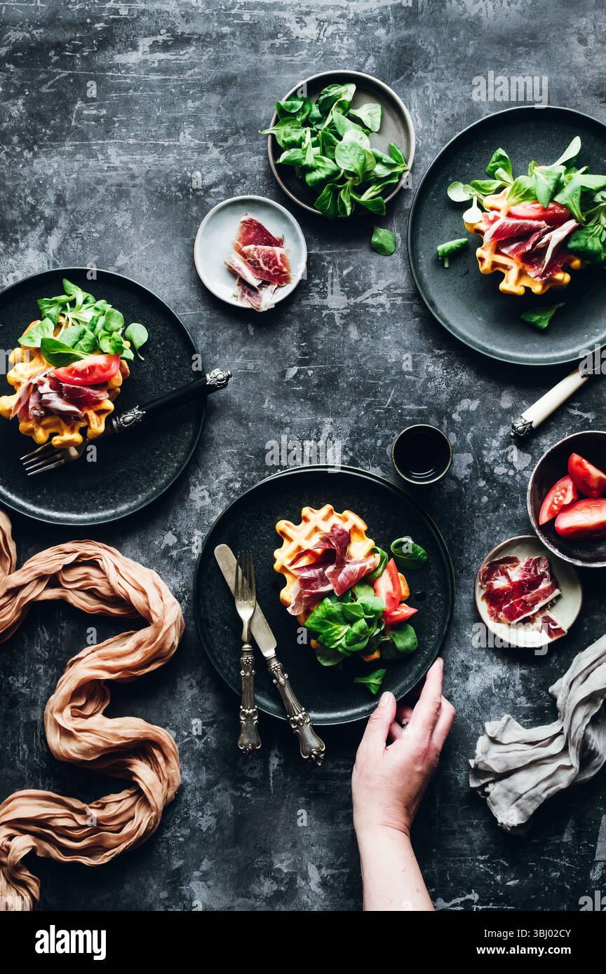 Elegante piatto gourmet con waffle di zucca splendidamente conditi con prosciutto iberico salato, verdure fresche e fette di pomodoro maturo, per creare un delizioso piatto Foto Stock
