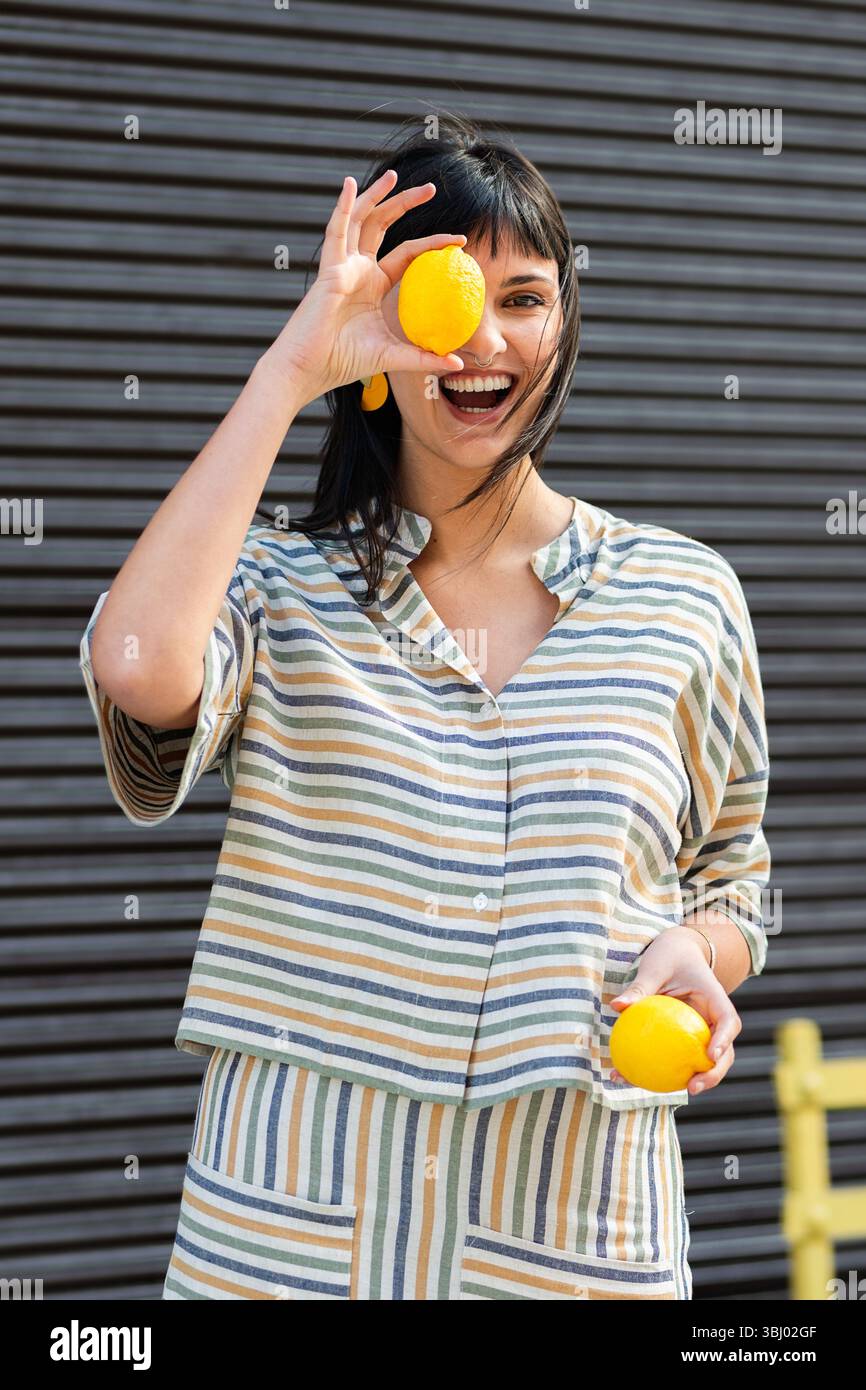 Donna gioiosa in abito a righe che regge limoni, simboleggia freschezza e felicità. Il suo sorriso trasmette positività e apprezzamento per la semplice natura Foto Stock
