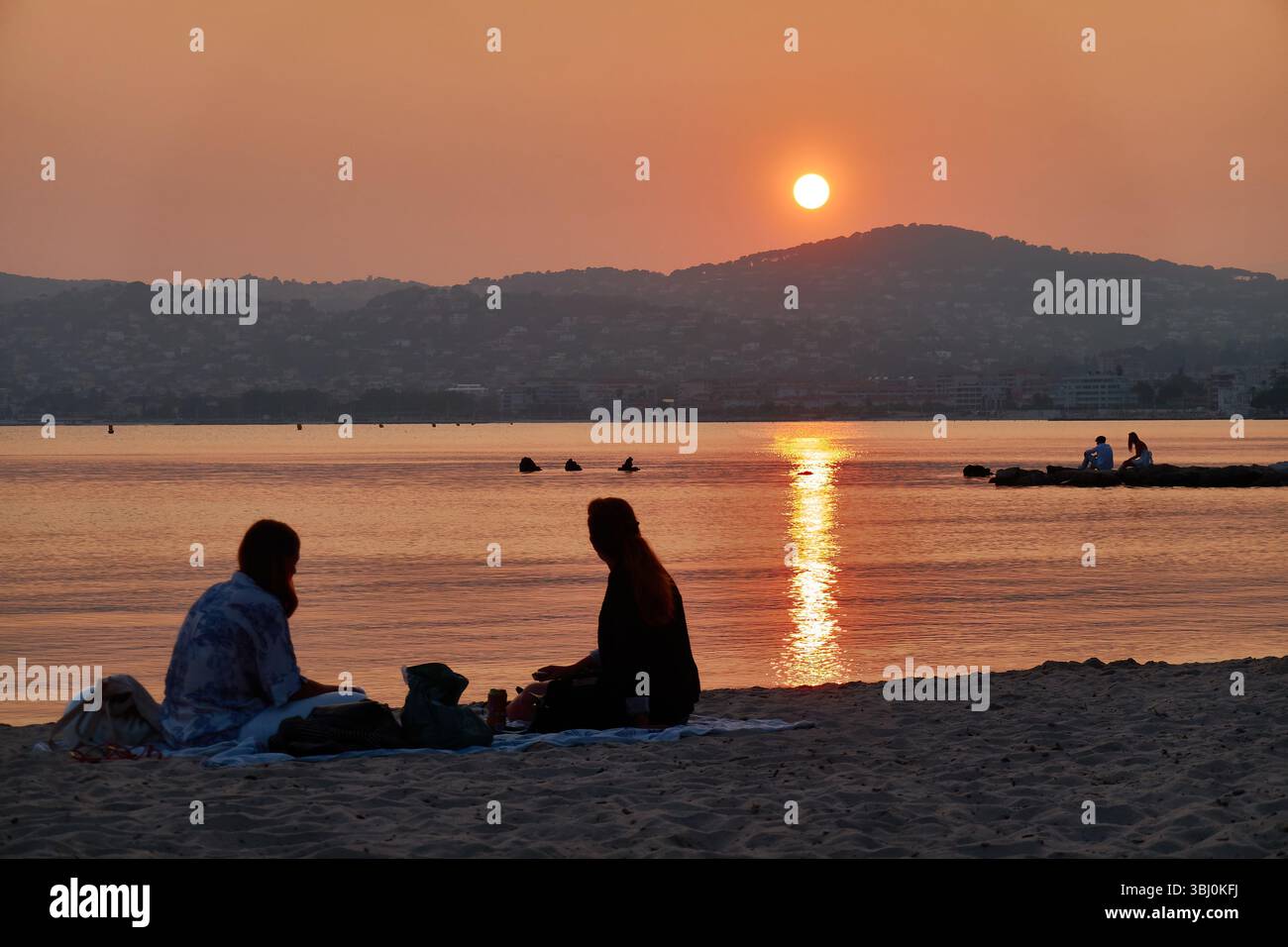 Amici sulla spiaggia che si godono un tranquillo tramonto che si riflette sul mare calmo con colline sullo sfondo. Foto Stock