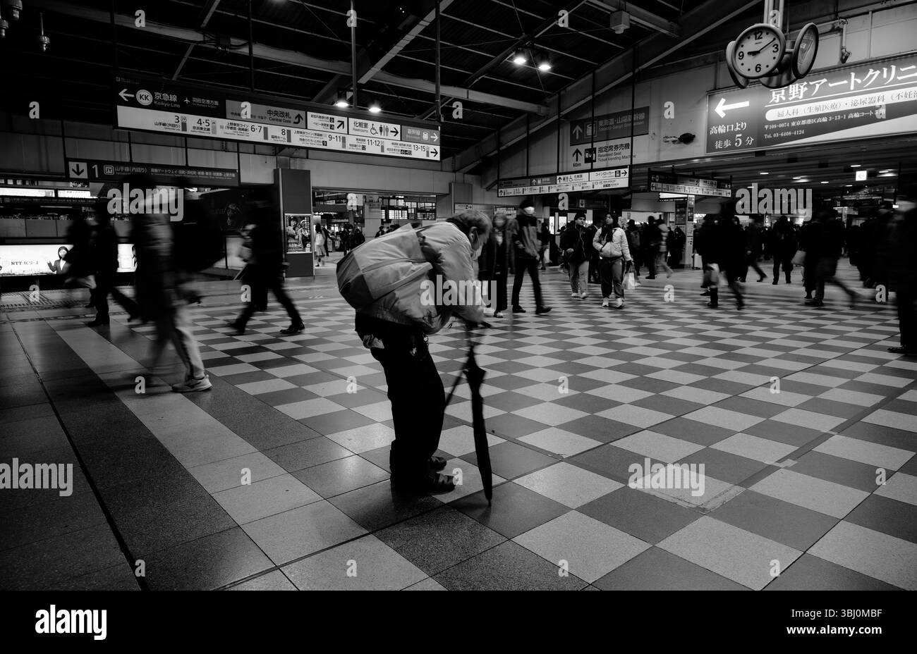 Un anziano cacciato si muove lentamente attraverso il pavimento a scacchi di una vivace stazione di Tokyo Foto Stock
