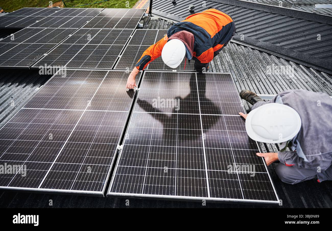 Tecnici professionisti che regolano i pannelli fotovoltaici. Installazione di impianti solari sul tetto della casa. Vista dall'alto di due lavoratori maschi in muri e tute da lavoro che fissano la batteria solare. Foto Stock