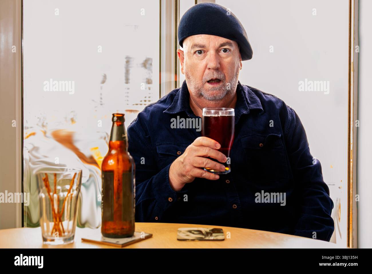 Un uomo si siede a un tavolo con una bottiglia di birra e bastoncini di pretzel, tenendo un bicchiere di birra scura con un'espressione sorpresa Foto Stock