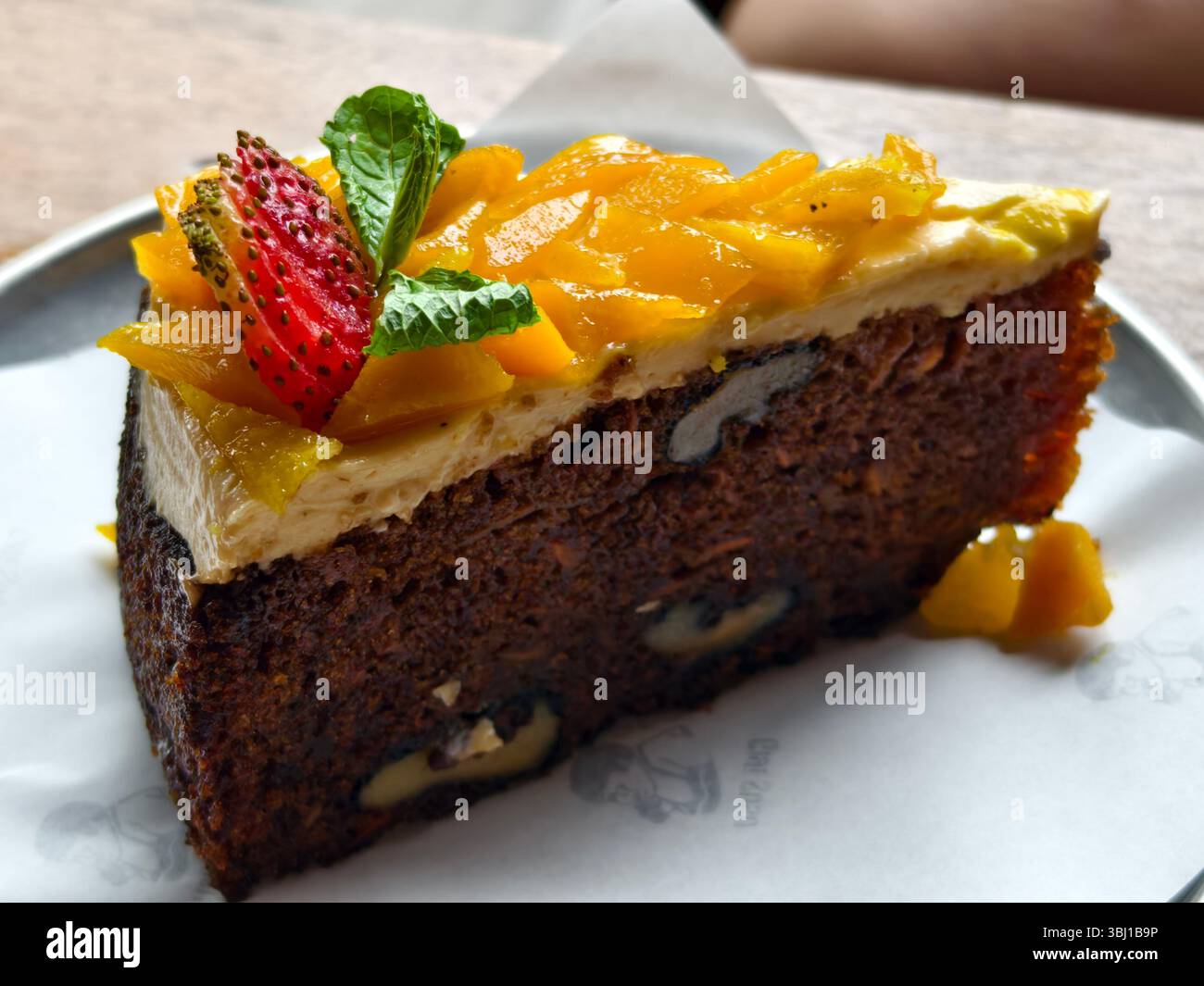 Una fetta di torta di carote con noci, ricoperta di glassa di formaggio spalmabile, mango a dadini, una fragola e un contorno di foglie di menta Foto Stock