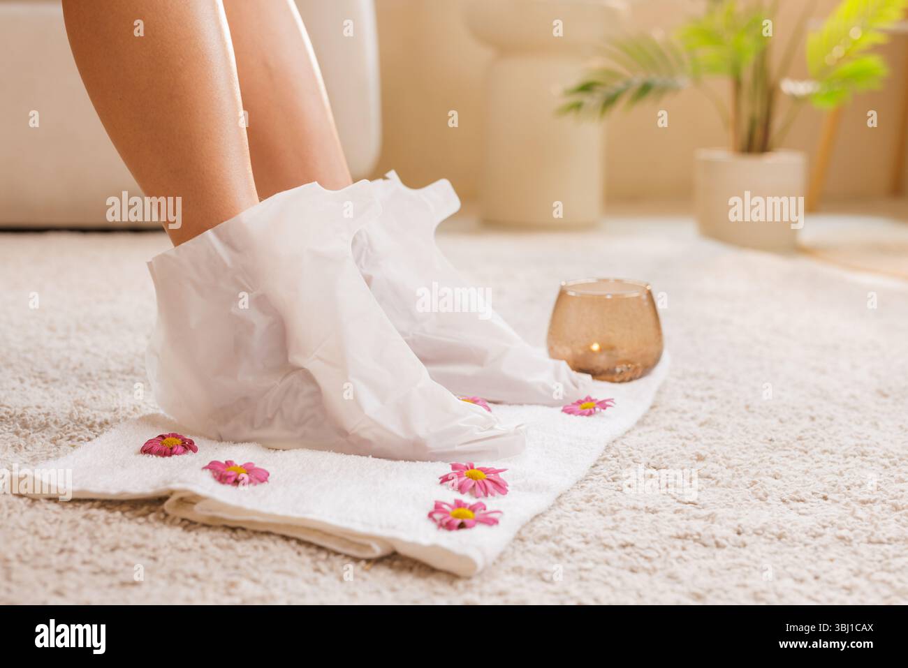 I piedi della donna in calze termali su un asciugamano fiorito con una candela, simboleggiano la cura dei piedi a casa, l'idratazione della pelle e la routine di auto-coccole. Foto Stock