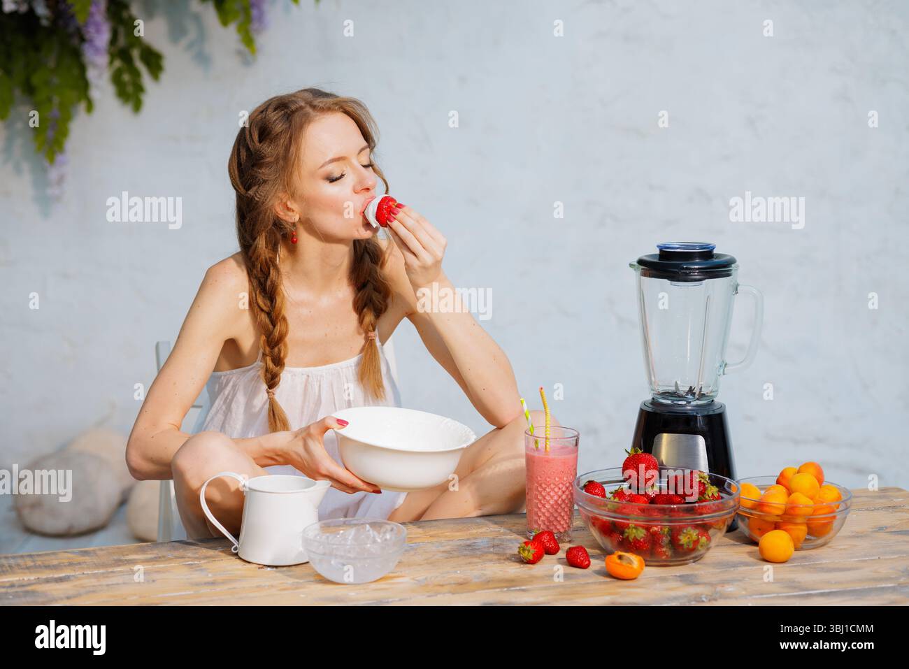 Donna che mangia fragole con panna montata al tavolo all'aperto con frullato, frullatore e ciotole di frutta fresca. Atmosfera da dessert estiva. Foto Stock