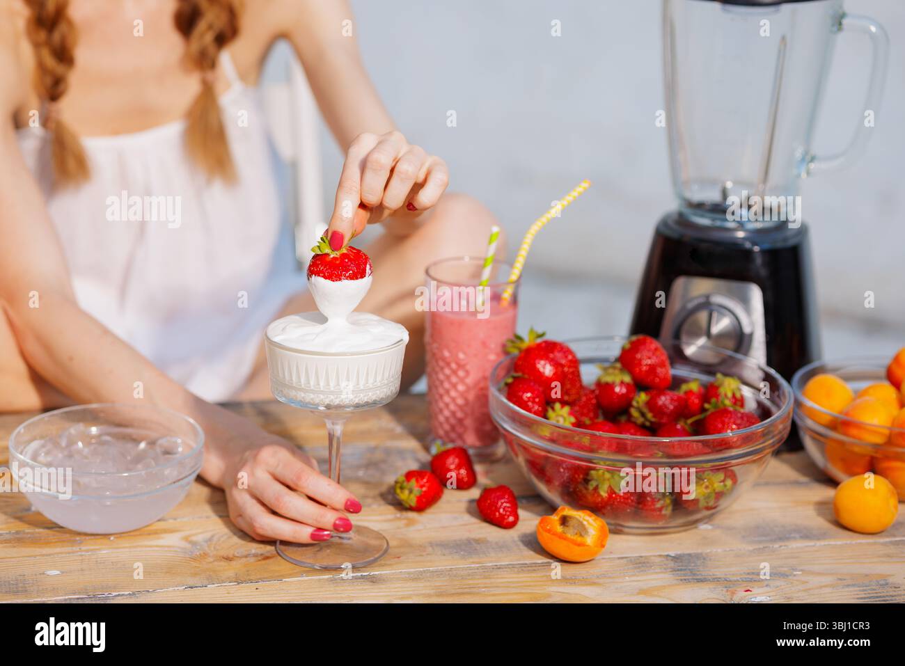 La donna immerge le fragole nella panna al tavolo estivo con frullato, frutta fresca e frullatore. Snack salutari. Foto Stock