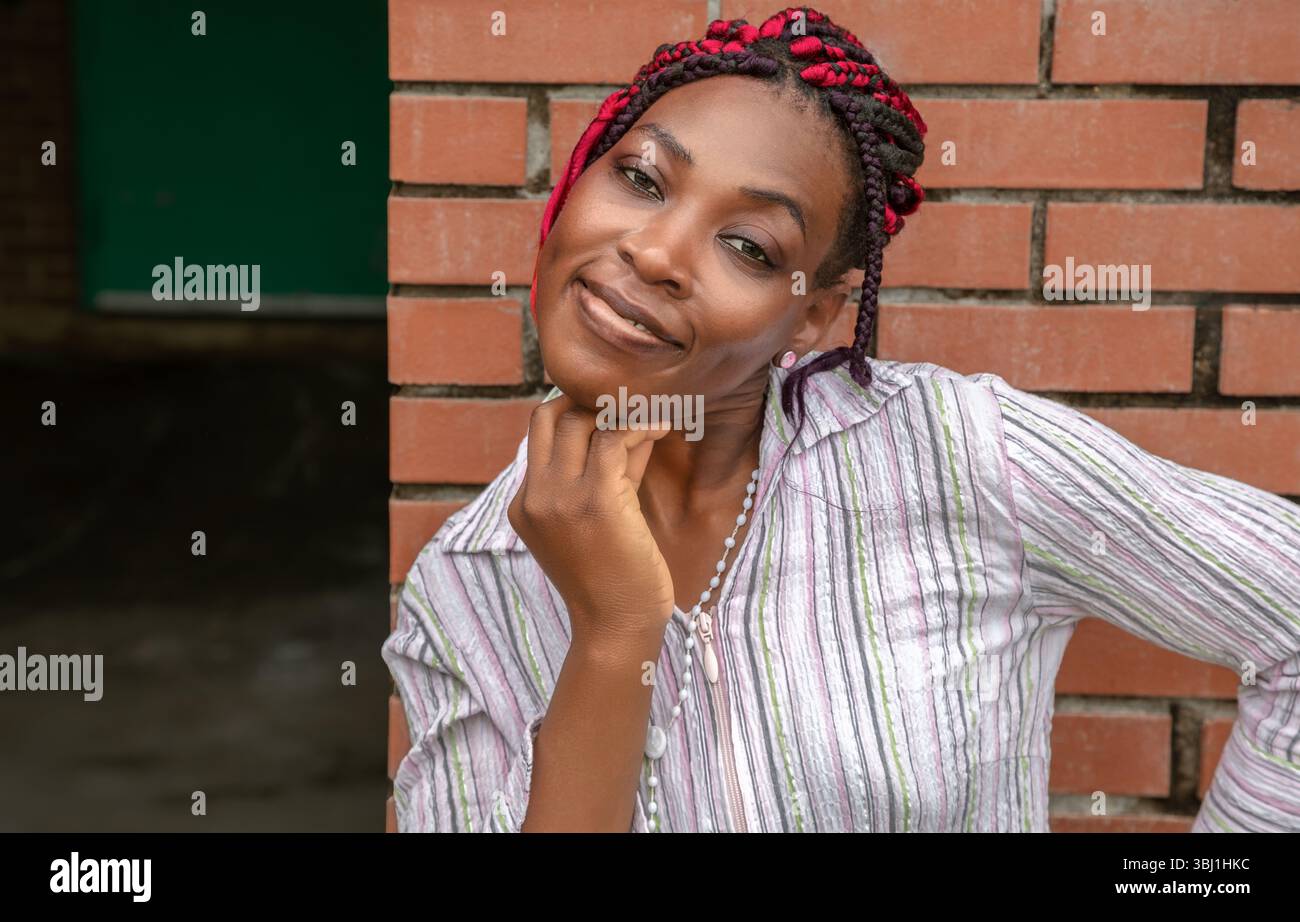 Allegra donna afroamericana con acconciatura intrecciata rossa che tocca il mento con cura. Ritratto stile di vita all'aperto in camicia a righe contro il muro di mattoni Foto Stock