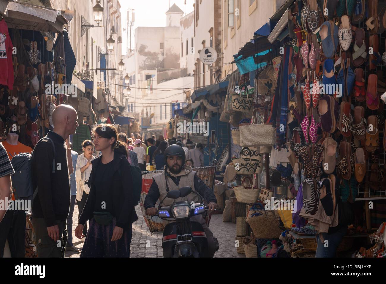 Essaouira, Marocco - 23 febbraio 2025: Un uomo locale guida uno scooter nelle strade affollate della medina di Essaouira, fiancheggiata da negozi di souvenir. Foto Stock