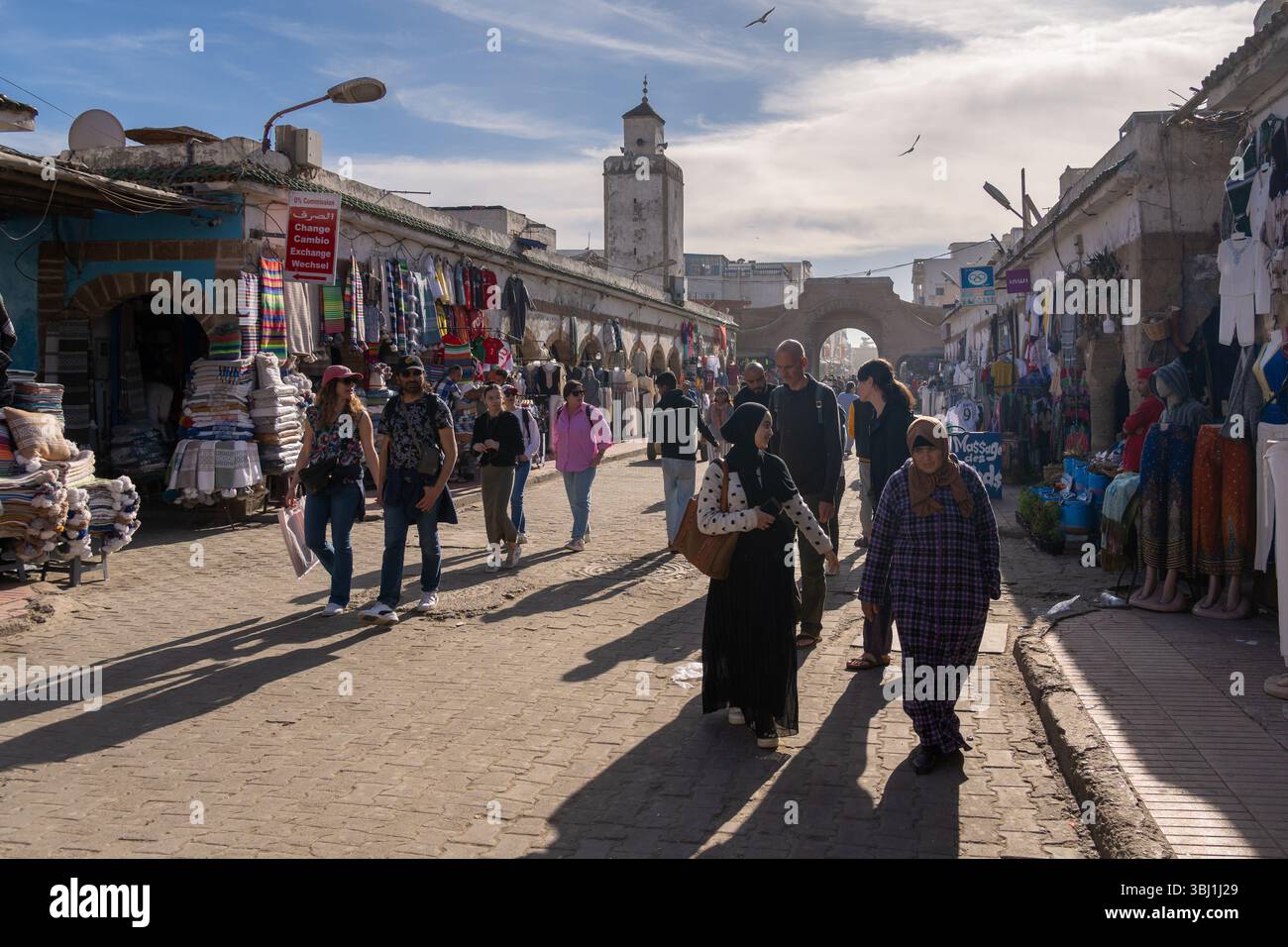 Essaouira, Marocco - 23 febbraio 2025: Donne e turisti locali camminano per le strade della medina di Essaouira con un minareto moschea nella città vecchia. Foto Stock