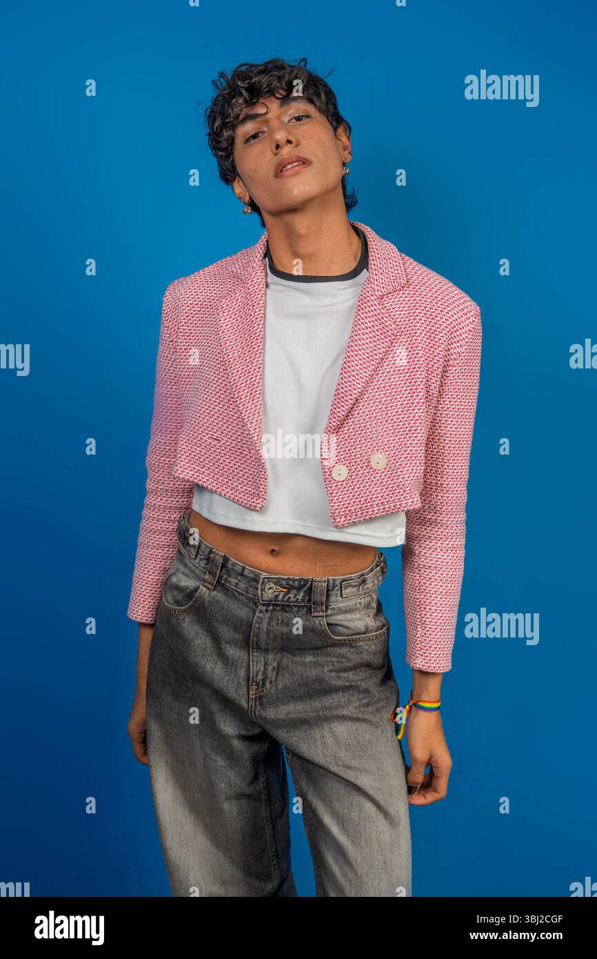 Giovane modello latino alla moda con giacca rosa, t-shirt bianca e jeans, in posa con bracciale lgbt su sfondo blu Foto Stock