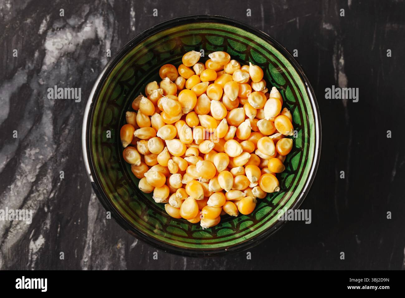 Essiccare i chicchi di mais in un recipiente verde. Grani organici su tavolo di marmo scuro. Ingrediente alimentare naturale. Cucina rustica. Semi testurizzati gialli. Foto Stock