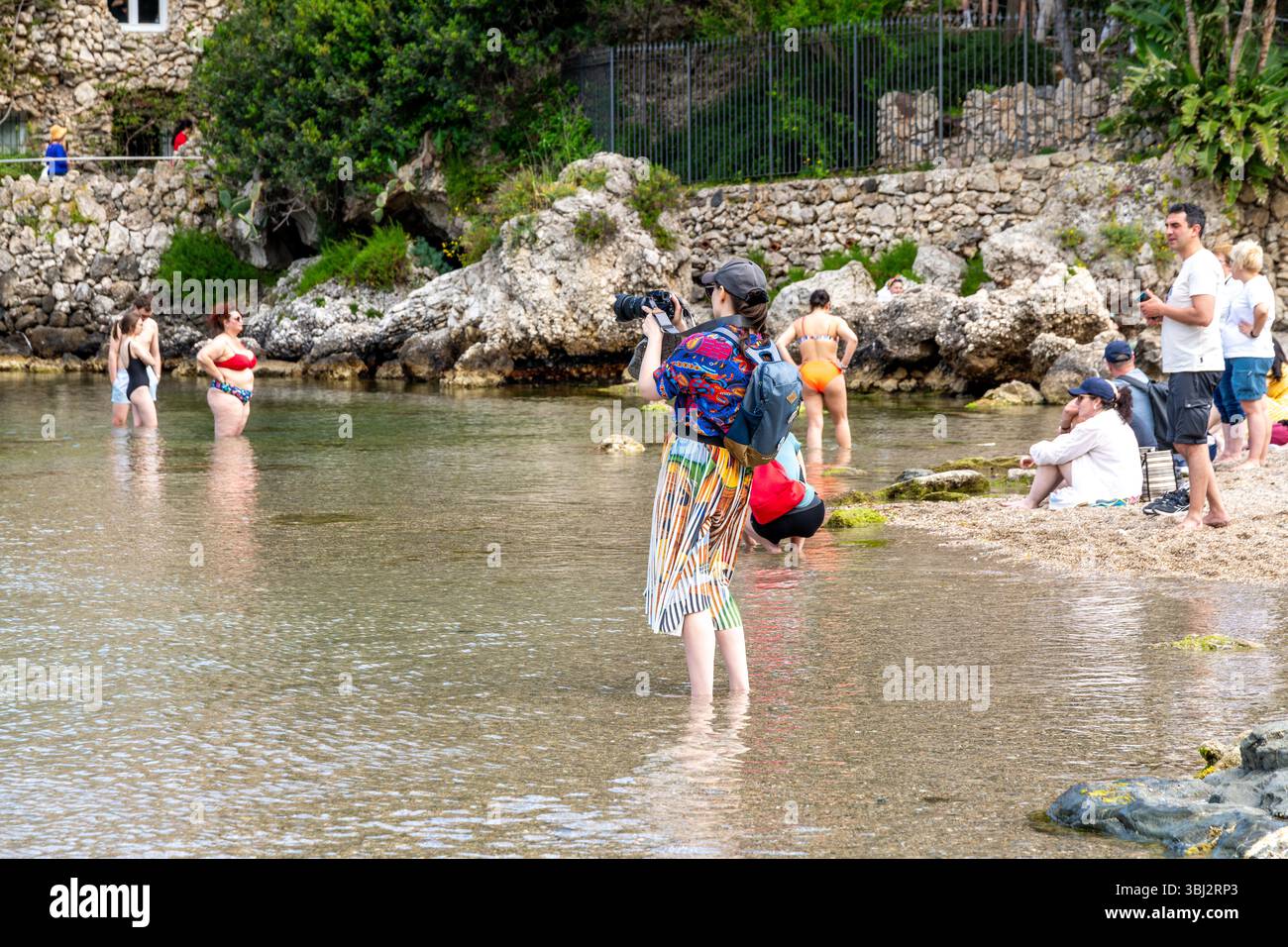 Taormina, Sizilien - Italia - 05-04-2025: Donna con fotocamera in piedi in acque poco profonde che scatta foto, circondata da turisti, costa rocciosa e vegetazione Foto Stock