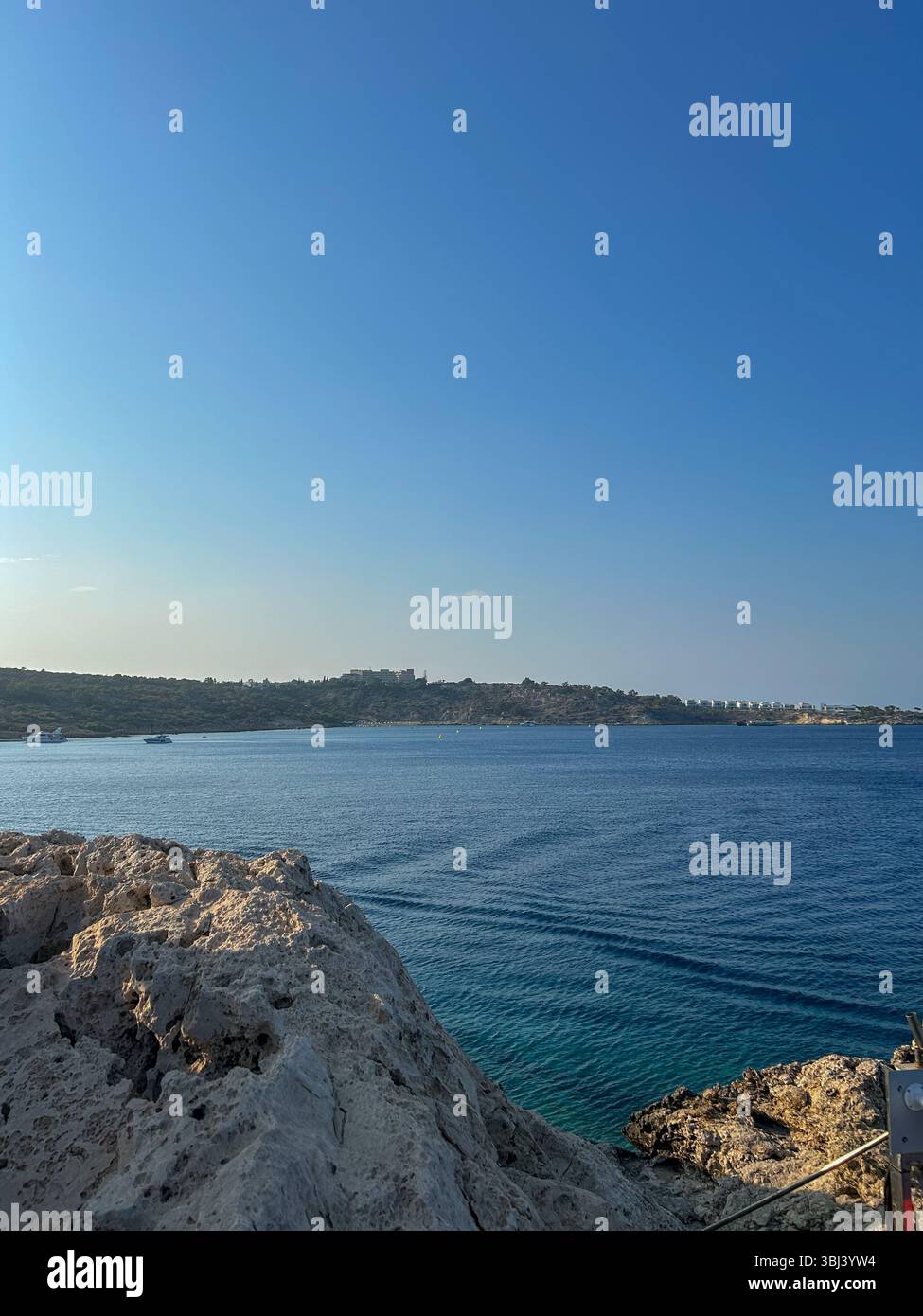 Vista panoramica di una costa rocciosa con mare blu calmo e cristallino e barche lontane sotto un cielo luminoso e senza nuvole, perfetto paesaggio costiero estivo. - Immagine stock catturata con smartphone