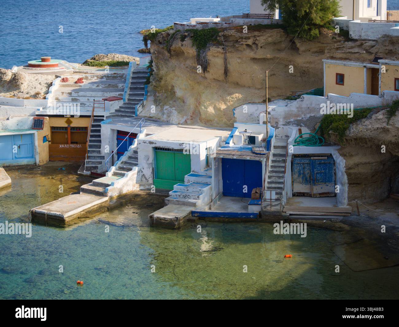 Colorate capanne di pesca nella Baia di Mandrakia sull'isola di Milos, con facciate bianche, porte vivaci, acqua turchese e scale che conducono giù dalla scogliera. Foto Stock