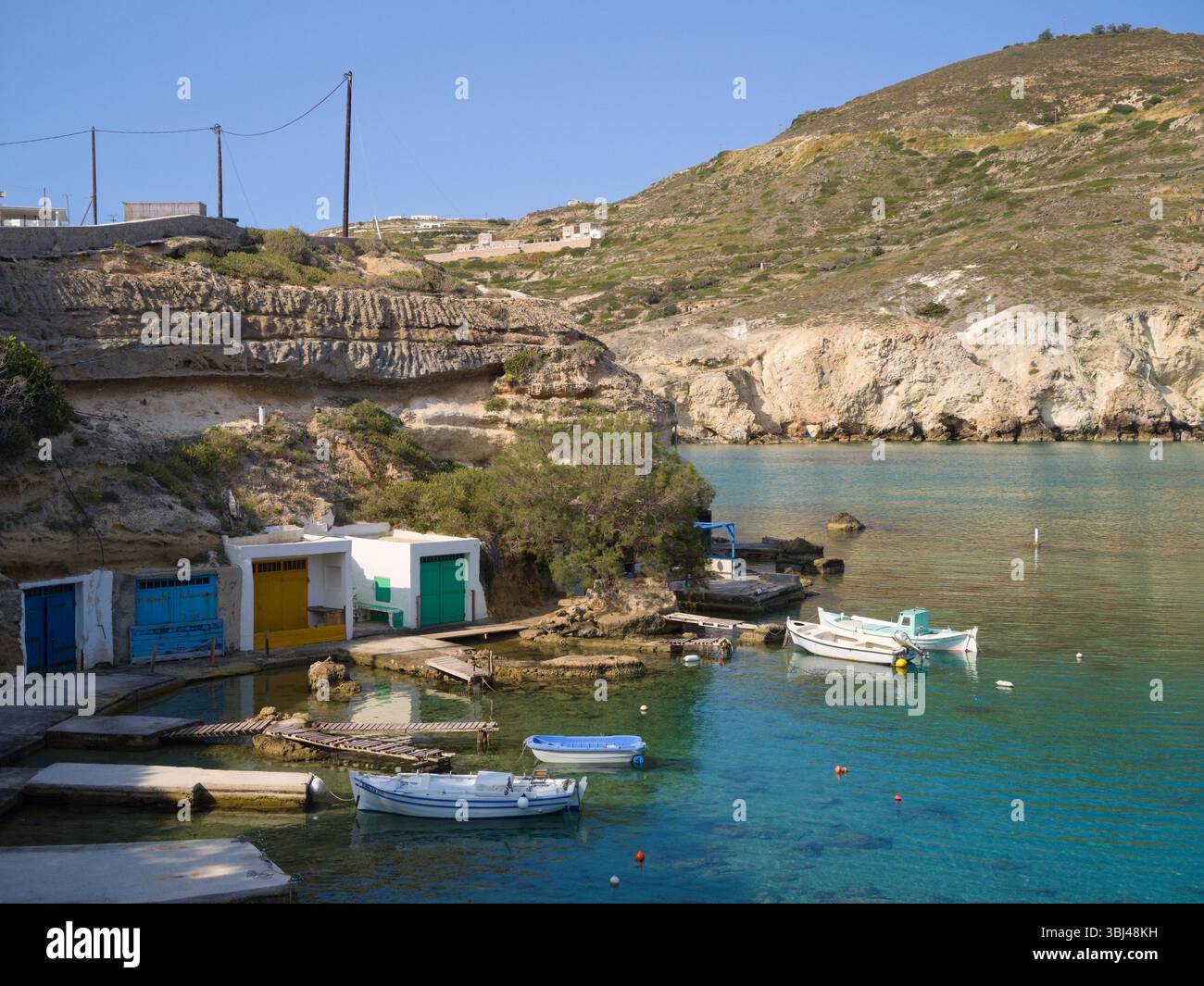 Vista panoramica delle capanne di pesca di Mandrakia con porte luminose lungo bianche scogliere, che si innalzano sopra le calme acque turchesi sotto un soleggiato cielo greco. Foto Stock
