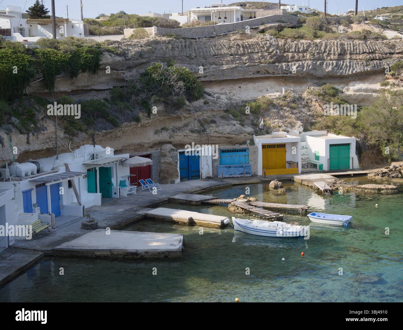Capanne da pesca con porte luminose fiancheggiano le bianche scogliere di Mandrakia, Milos. L'acqua turchese e la luce del sole creano una serena vista del Mediterraneo. Foto Stock