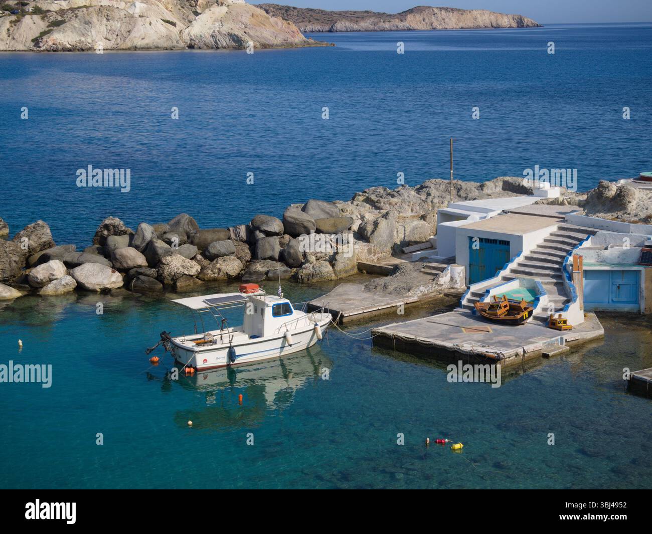 Piccola barca su lungo molo in pietra nella baia di Mandrakia, Milos, con alcune capanne bianche e il mare Egeo aperto sotto cieli limpidi e soleggiati. Foto Stock