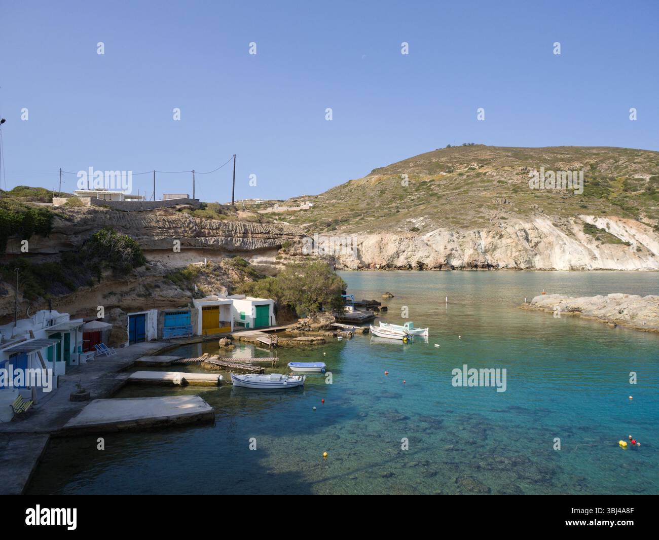Le bianche scogliere sovrastano le vivaci capanne del porto di Mandrakia e la baia turchese in questa vista soleggiata da Milos, Grecia. Foto Stock