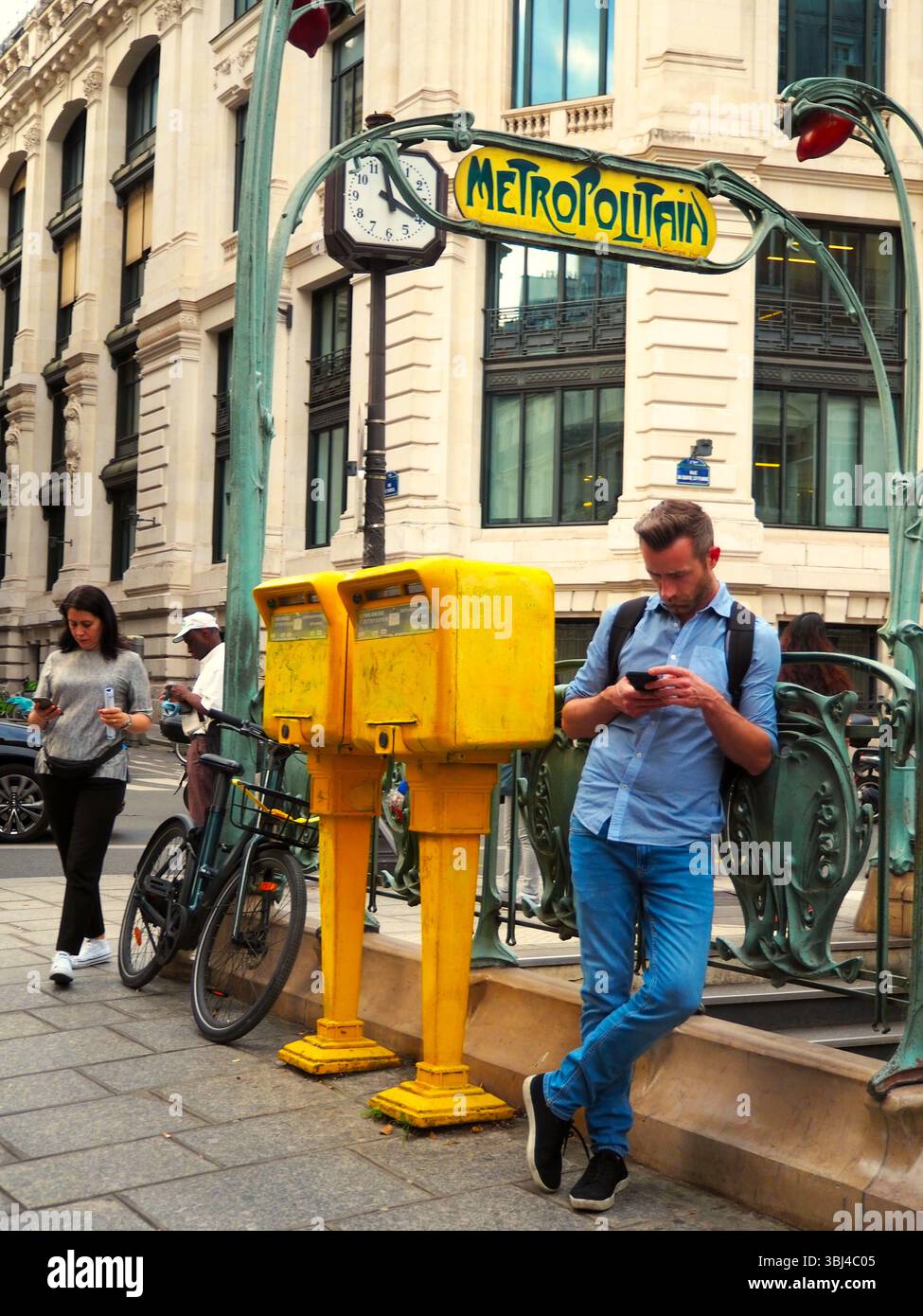 Uomo che utilizza uno smartphone vicino alle cassette postali gialle all'ingresso art nouveau della metropolitana di Parigi Foto Stock
