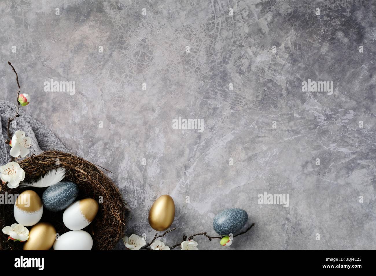 Eleganti uova di Pasqua in un nido, circondate da delicati fiori bianchi su sfondo grigio testurizzato. Design del biglietto di auguri di buona Pasqua. Foto Stock