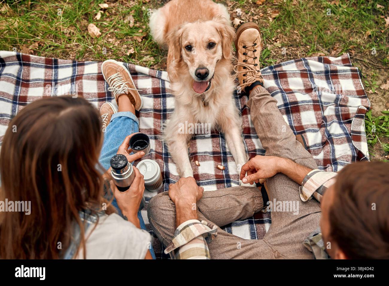 Una coppia allegra gode di un delizioso picnic su una spaziosa coperta con il loro giocoso Golden retriever in mezzo allo splendido e sereno sfondo di un l Foto Stock