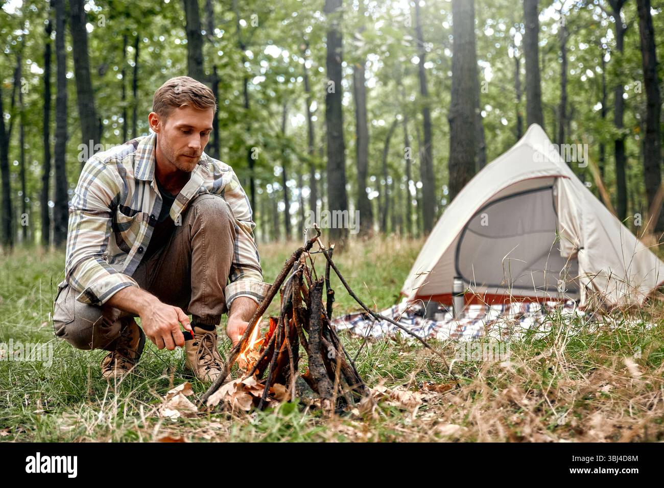 Un uomo sta preparando sapientemente un fuoco caldo e invitante nella tranquilla e tranquilla foresta, con una tenda accogliente allestita nelle vicinanze che abbraccia completamente il tranqui Foto Stock