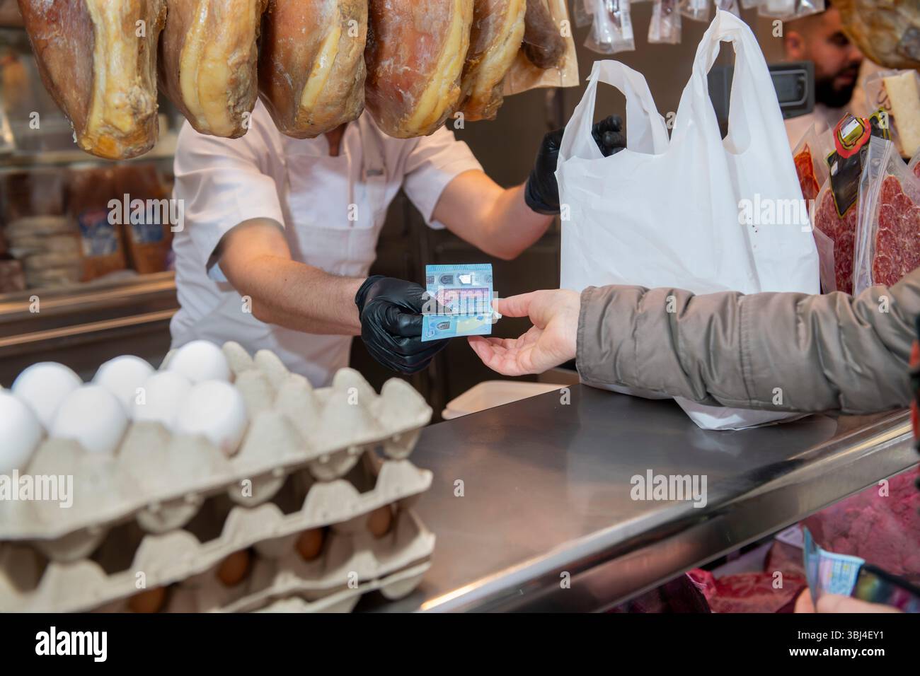 Macellaio che indossa guanti neri che ricevono banconote in euro dal cliente che acquista generi alimentari in macelleria con le gambe di prosciutto stagionato appese Foto Stock