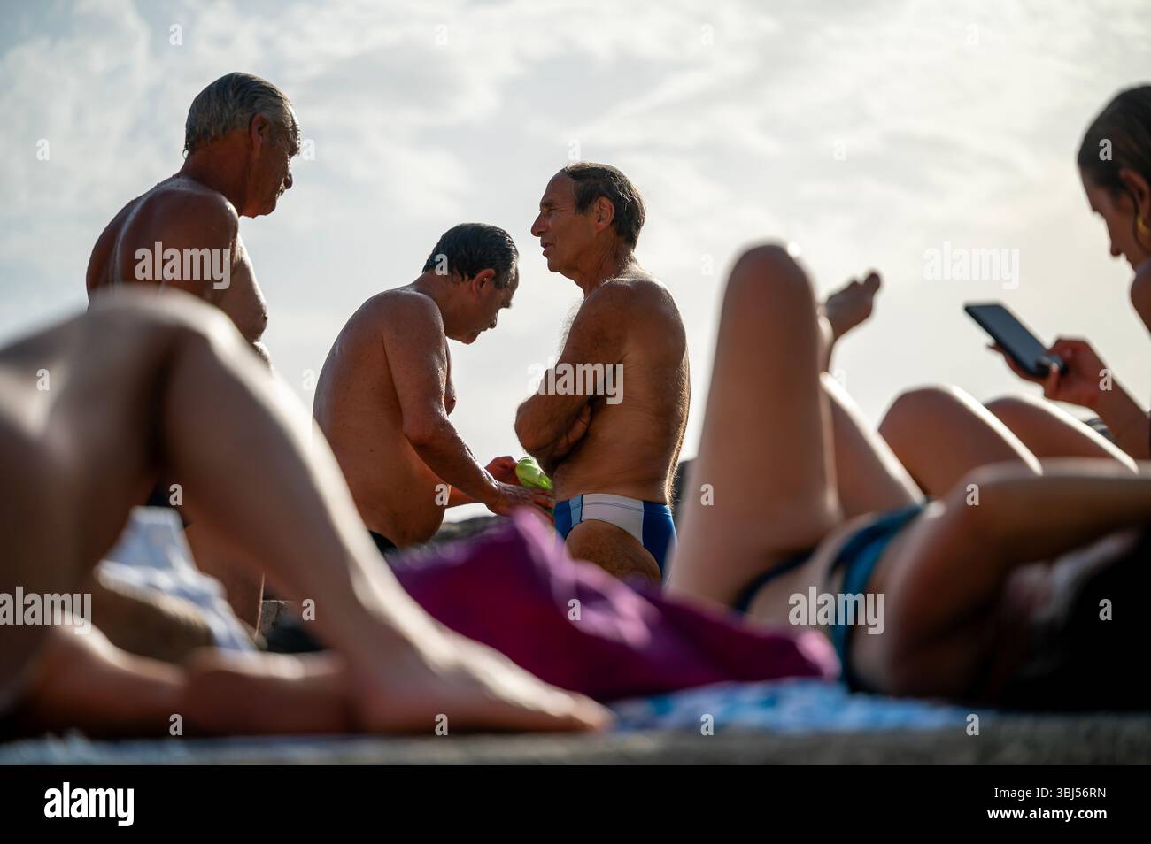 Tre uomini italiani più anziani si trovano sulla spiaggia di Vernazza, in Italia, incorniciati umoristicamente tra le gambe delle donne che prendono il sole in primo piano. Foto Stock
