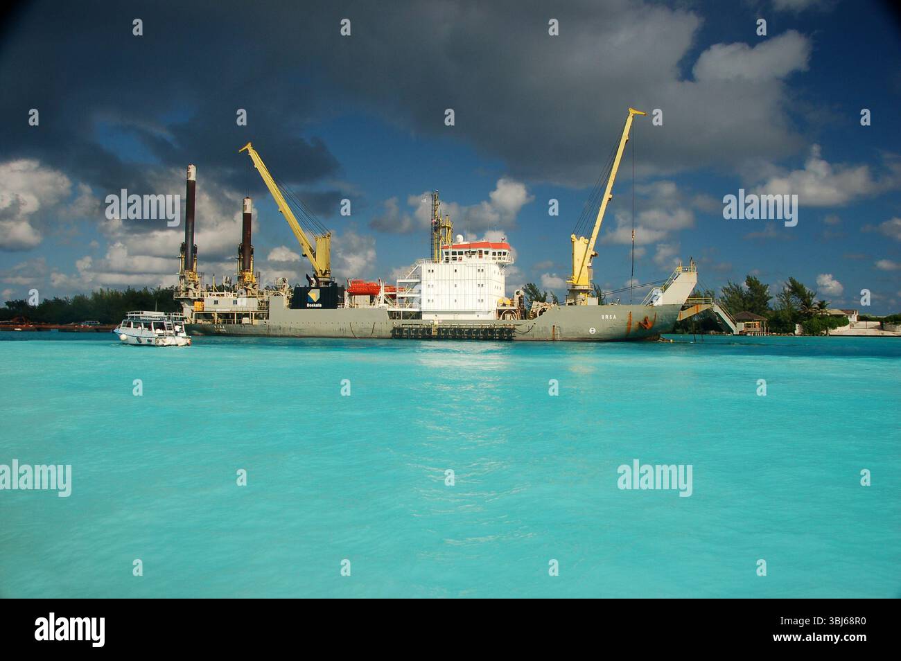 Nave da carico industriale con grandi gru ancorata in acque turchesi sotto un cielo spettacolare in un porto caraibico, con una piccola barca nelle vicinanze Foto Stock