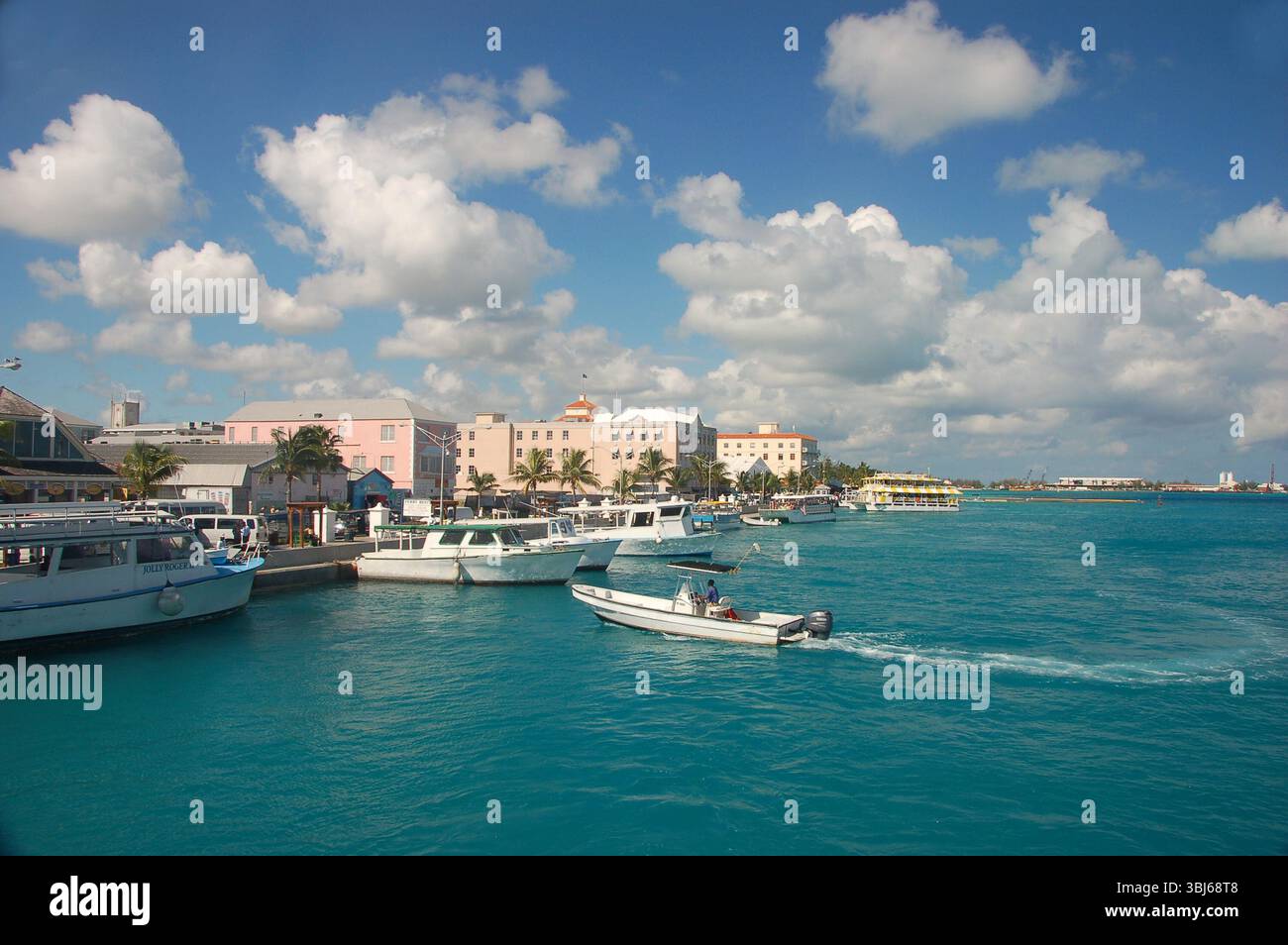 Le barche attraccavano lungo il vivace lungomare di Nassau, Bahamas, con edifici color pastello, palme e un motoscafo che attraversa le acque turchesi Foto Stock