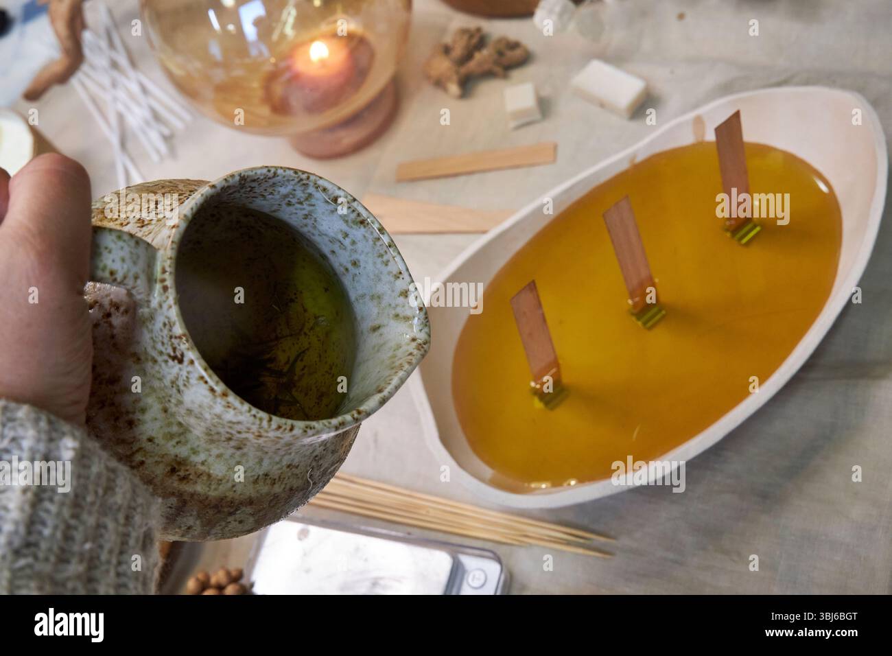 Primo piano del processo di preparazione delle candele: La cera fusa viene versata da una caraffa in un recipiente di ceramica con stoppini di legno. Una scena calda e artigianale Foto Stock