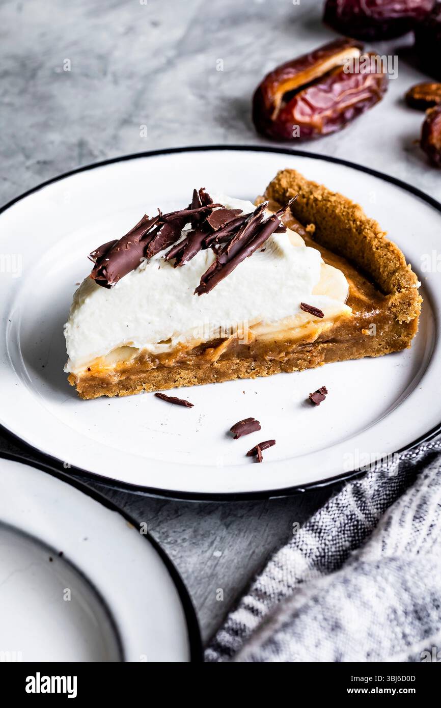 Una fetta di torta condita con panna montata e scaglie di cioccolato su un piatto bianco, con datteri sullo sfondo. Foto Stock