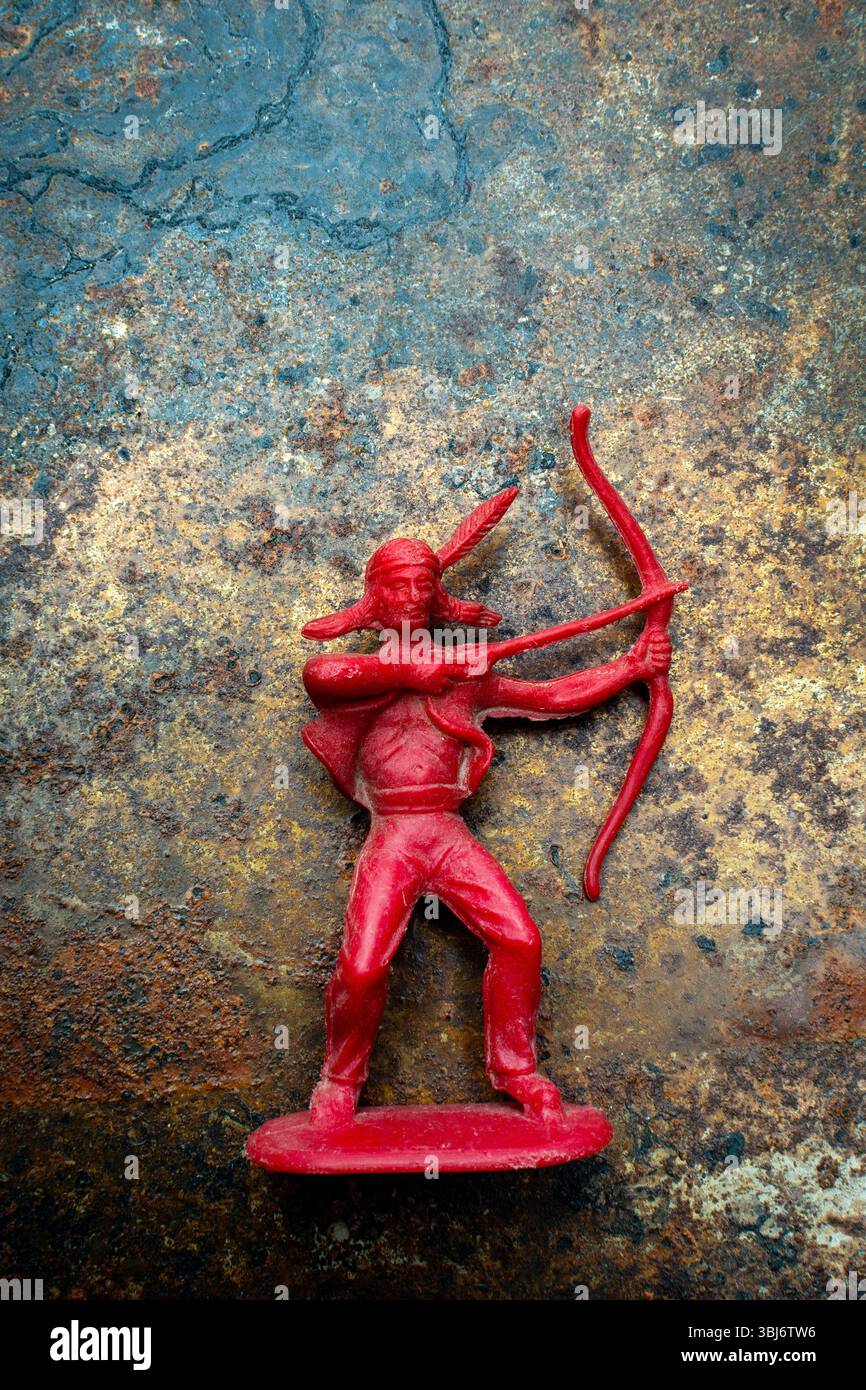 Un giocattolo di plastica rosso archer rappresenta una figura dei nativi americani, posata con un fiocco e una freccia su una superficie ruvida e ruggine. T Foto Stock Un giocattolo di plastica rosso archer rappresenta una figura dei nativi americani, posata con un fiocco e una freccia su una superficie ruvida e ruggine. T Foto Stock