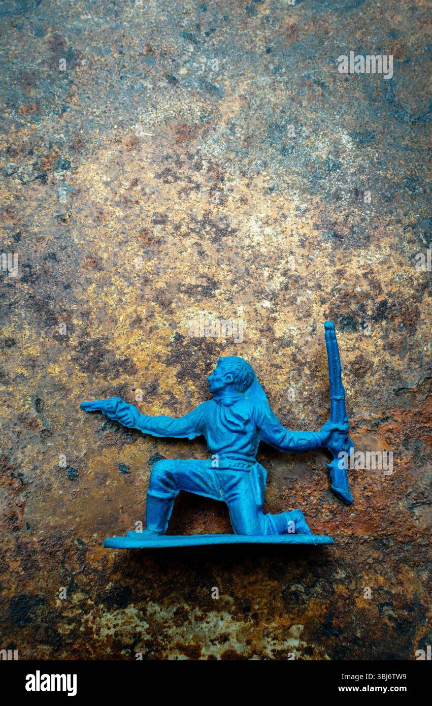 Un cowboy di plastica blu si inginocchia, tenendo una pistola in una mano e un fucile nell'altra, posizionato su una superficie metallica testurizzata Foto Stock