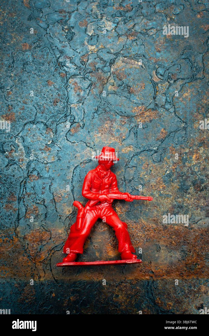 Un piccolo cowboy rosso di plastica si appoggia saldamente su uno sfondo rustico blu e marrone. La figura è dotata di un'arma Foto Stock